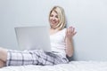 A young woman sits on a bed with a laptop and chats online. Beautiful blonde in pajamas against a gray wall. Remote work and