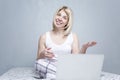 A young woman sits on a bed with a laptop and chats online. Beautiful blonde in pajamas against a gray wall. Remote work and