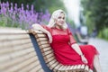 A young woman sits on a bench in a blooming park. Smiling beautiful blonde in a bright red dress