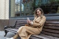 A young woman sits on a bench in the city with a phone in her hands. Beautiful stylish blonde in a beige coat. Remote work and Royalty Free Stock Photo