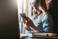 Young woman sits in cafe on windowsill and uses smartphone. Hipster girl checking e-mail, chatting, blogging, working. Royalty Free Stock Photo