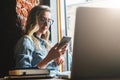 Young woman sits in cafe on windowsill and uses smartphone. Hipster girl checking e-mail, chatting, blogging, working. Royalty Free Stock Photo