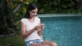 Young woman sits on edge of pool and uses phone for work, greneery on background