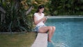 Young woman sits on edge of pool and uses phone for work, greneery on background