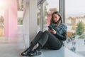 Woman sits on floor near window, uses smartphone.Hipster girl looking on screen of phone,blogging, browsing internet.