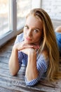 A young woman sits in front of the window on a large wooden sill. The girl is sitting on the windowsill. A young blonde is sitting Royalty Free Stock Photo