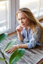 A young woman sits in front of the window on a large wooden sill. The girl is sitting on the windowsill. A young blonde is sitting Royalty Free Stock Photo