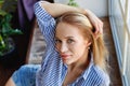 A young woman sits in front of the window on a large wooden sill. The girl is sitting on the windowsill. A young blonde is sitting Royalty Free Stock Photo