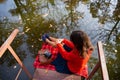 A young woman sits with her back on a bridge near a lake on a sunny day. Plaid, camera, suitcase Royalty Free Stock Photo