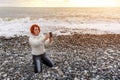 Young woman sits on her lap on shingly shore against the backdrop of the sea and sunset and takes a selfie Royalty Free Stock Photo