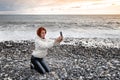 Young woman sits on her lap on shingly shore against the backdrop of the sea and sunset and takes a selfie Royalty Free Stock Photo