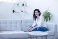 Young woman sits on her sofa in modern apartment and types on laptop with a smile on her face