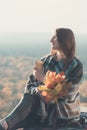 Young woman with sits on a hill. Paper cup in hands and a bouquet of yellow leaves. Enjoying a sunny day