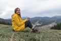 Young woman sits on hillside, drinking tea and smiling. Portrait of tourist girl in yellow jacket on the mountains Royalty Free Stock Photo