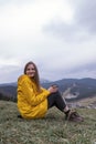 Young woman sits on hillside, drinking tea and smiling. Portrait of tourist girl in yellow jacket on the mountains. Vertical frame Royalty Free Stock Photo