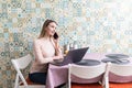 Young woman sits at the kitchen table using a laptop and talking on a cell phone Royalty Free Stock Photo