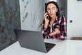 Young woman sits at the kitchen table using a laptop and talking on a phone Royalty Free Stock Photo