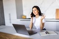 Young woman sits at the kitchen table using a laptop and talking on a phone Royalty Free Stock Photo