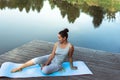 A young woman sits on a mat in a free pose by the lake. Yoga class in nature