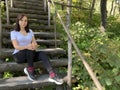 Young woman sits on old wooden staircase in woodland. Adult brunette poses, looking at camera and resting on stairs in Royalty Free Stock Photo