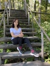 Young woman sits on old wooden staircase in woodland. Adult brunette poses, looking at camera and resting on stairs in Royalty Free Stock Photo