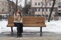 Young woman sits on park bench in city and talks on phone . Girl in beige sheepskin coat rests outside in winter