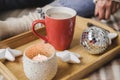 Young woman sits on plaid in cozy knitted woolen white sweater and holds cup of cocoa in her hands. Hygge wooden tray