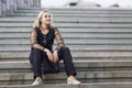 Young woman sits on the steps in the street. Smiling blonde in black jeans