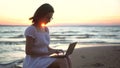 A young woman sits on a stone on the beach by the sea with a laptop in her hands. A girl in a white dress at sunset is Royalty Free Stock Photo