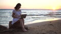 A young woman sits on a stone on the beach by the sea with a laptop in her hands. A girl in a white dress at sunset is Royalty Free Stock Photo