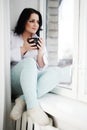 Young woman sits on window sill with cup in hands and looks out of window. Morning of winter.