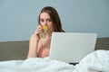 Young woman sitting in bed with laptop computer and drinking water with lemon