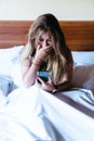 Young woman sitting on bed looking at her smartphone with a worried, shocked and surprised expression while covering her mouth Royalty Free Stock Photo