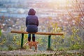 Young woman sitting on bench in autumn city park and looking at cityscape, back view