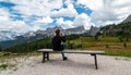 Young woman sitting at bench, drinking glass of red wine, looking at Cima Ambrizzola in front of Rifugio Cinque Torri. Dolomites,