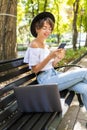 Young woman sitting on bench in green park on summer day and reading text message on cell phone while using silver laptop Royalty Free Stock Photo