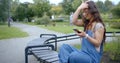 Young woman is sitting on bench in park, reading and typing messages on smartphone Royalty Free Stock Photo