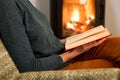 Young woman is sitting on a chair near a fireplace and reading a book, holding a cup of tea Royalty Free Stock Photo