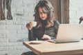 Young woman sitting in coffee shop at wooden table, drinking coffee and using smartphone.On table is laptop. Girl browsing interne Royalty Free Stock Photo