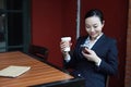 Young woman sitting in coffee shop at wooden table, drinking coffee and using smartphone.On table is laptop Royalty Free Stock Photo