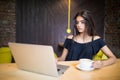 Young woman sitting in coffee shop at wooden table, drinking coffee and using smartphone. On table is laptop. Girl browsing intern Royalty Free Stock Photo