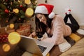 Young woman sitting with cute cat and looking at laptop under christmas tree with presents. Happy girl in santa hat chatting Royalty Free Stock Photo