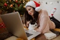 Young woman sitting with cute cat and looking at laptop under christmas tree with presents. Happy girl in santa hat chatting Royalty Free Stock Photo