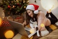 Young woman sitting with cute cat and looking at laptop under christmas tree with presents. Happy girl in santa hat chatting Royalty Free Stock Photo