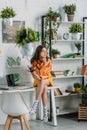 Young woman sitting on desk in room decorated with green plants and paintings on wall