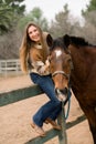 Young woman sitting on a fence while petting her brown horse Royalty Free Stock Photo