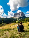 Young woman sitting in grass and looking at Sassolungo Langkofel mountain from Selva. Gardena Pass, Trentino Alto Adige, Italy.