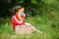 Young woman sitting on a grass with vintage suitcase, holding rainbow fan LGBT symbol, covered her face