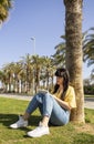 Young woman sitting on grass and writing in a notebook outside surrounding with palm trees.