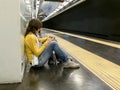 Young woman sitting on ground using phone while leaning on underground platform wall